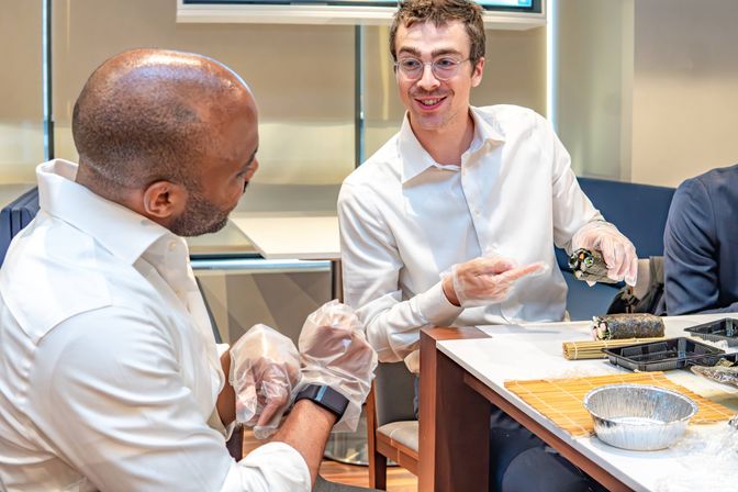 Two colleagues in white shirts making hand-rolled sushi at a conference table, wearing disposable gloves with bamboo mat, trays and sushi rolls visible — office team-building sushi workshop