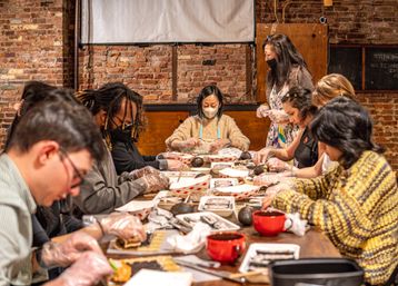 Group sushi-making class in a cozy brick-walled studio, participants wearing gloves and masks rolling sushi on bamboo mats around a communal table as an instructor looks on.