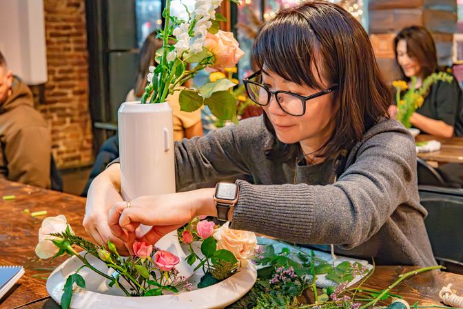 Person with glasses arranging pink and peach roses and greenery in a white vase on a wooden table during a cozy indoor floral workshop