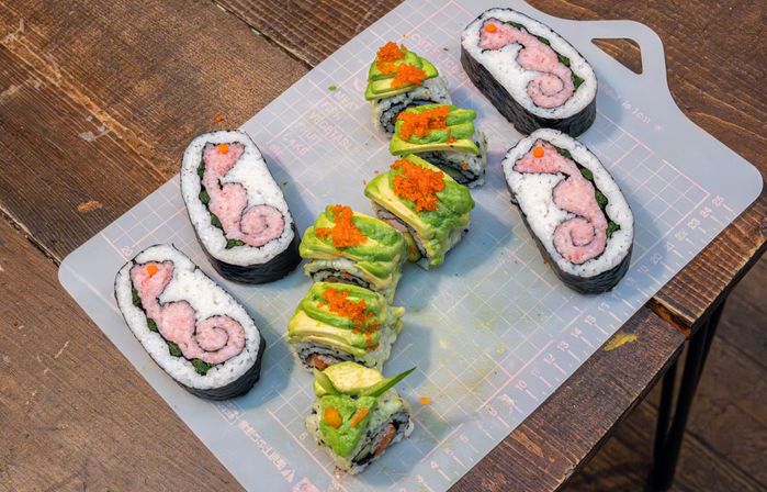 Assorted sushi rolls on a silicone cutting mat atop a rustic wooden table — avocado-topped maki with orange tobiko and spiral pink tuna-filled nori rolls.