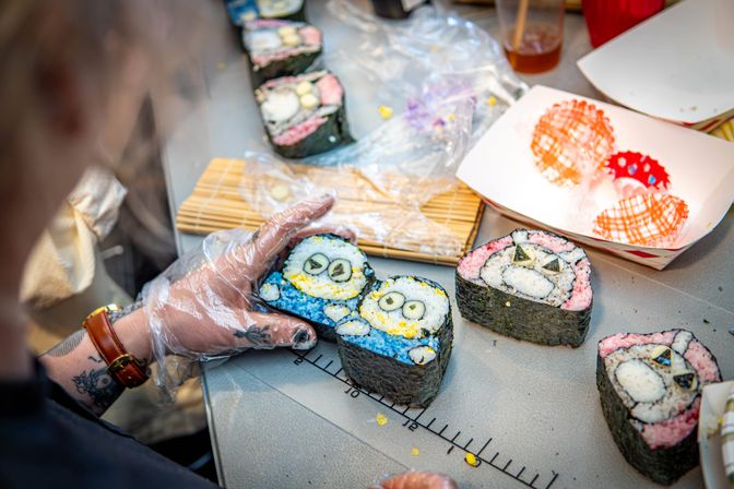 Hands in plastic gloves shaping colorful character sushi rolls with blue and pink rice and nori on a prep table with a bamboo mat and paper trays.