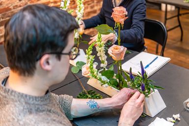 Person arranging peach roses, white snapdragons and purple spiky blooms in a white rectangular vase at an indoor floral-arranging workshop with exposed brick wall and notebook on the table.