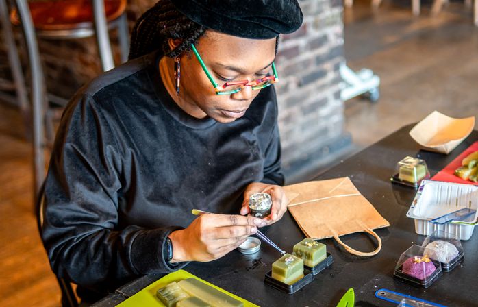Artist in a beret and colorful glasses hand-decorating small square green handmade soaps or confections with tweezers at an indoor craft studio table with packaging and tools.
