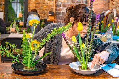 Participant arranging yellow tulips and daisy-like blooms with green foliage and purple accents in shallow bowls on a wooden table at a brick-walled flower-arranging workshop.