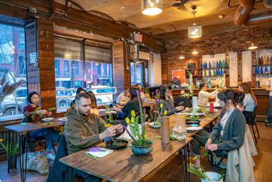 Cozy urban flower-arranging workshop in a brick-walled studio, people at wooden tables crafting tulip and greenery arrangements by large street-facing windows with colorful bottles on shelves.