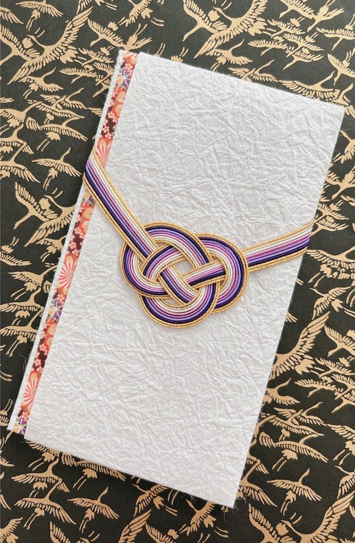 White textured washi envelope wrapped with a purple, lavender and gold mizuhiki knot and floral edge trim, photographed on black paper with a gold crane pattern