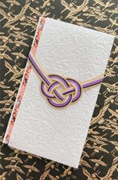 White textured washi envelope wrapped with a purple, lavender and gold mizuhiki knot and floral edge trim, photographed on black paper with a gold crane pattern