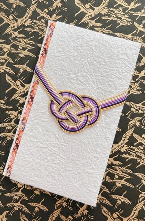 White textured washi envelope wrapped with a purple, lavender and gold mizuhiki knot and floral edge trim, photographed on black paper with a gold crane pattern