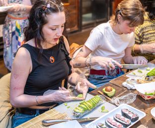 Two participants at an indoor sushi-making class assembling and slicing avocado-covered sushi rolls on a wooden table with bamboo rolling mats, avocado halves, and plated maki pieces.