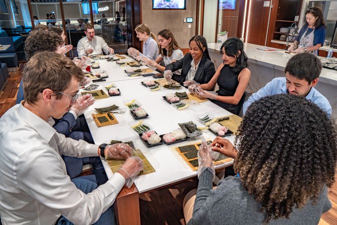 Team-building sushi-making class in a modern office kitchen — colleagues around a long table wearing gloves and using bamboo mats, nori and rice to roll sushi.