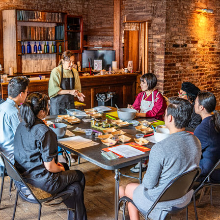 Cozy brick‑walled cooking workshop with an instructor demonstrating to a small group gathered around a table of bowls, ingredients, and recipe sheets.