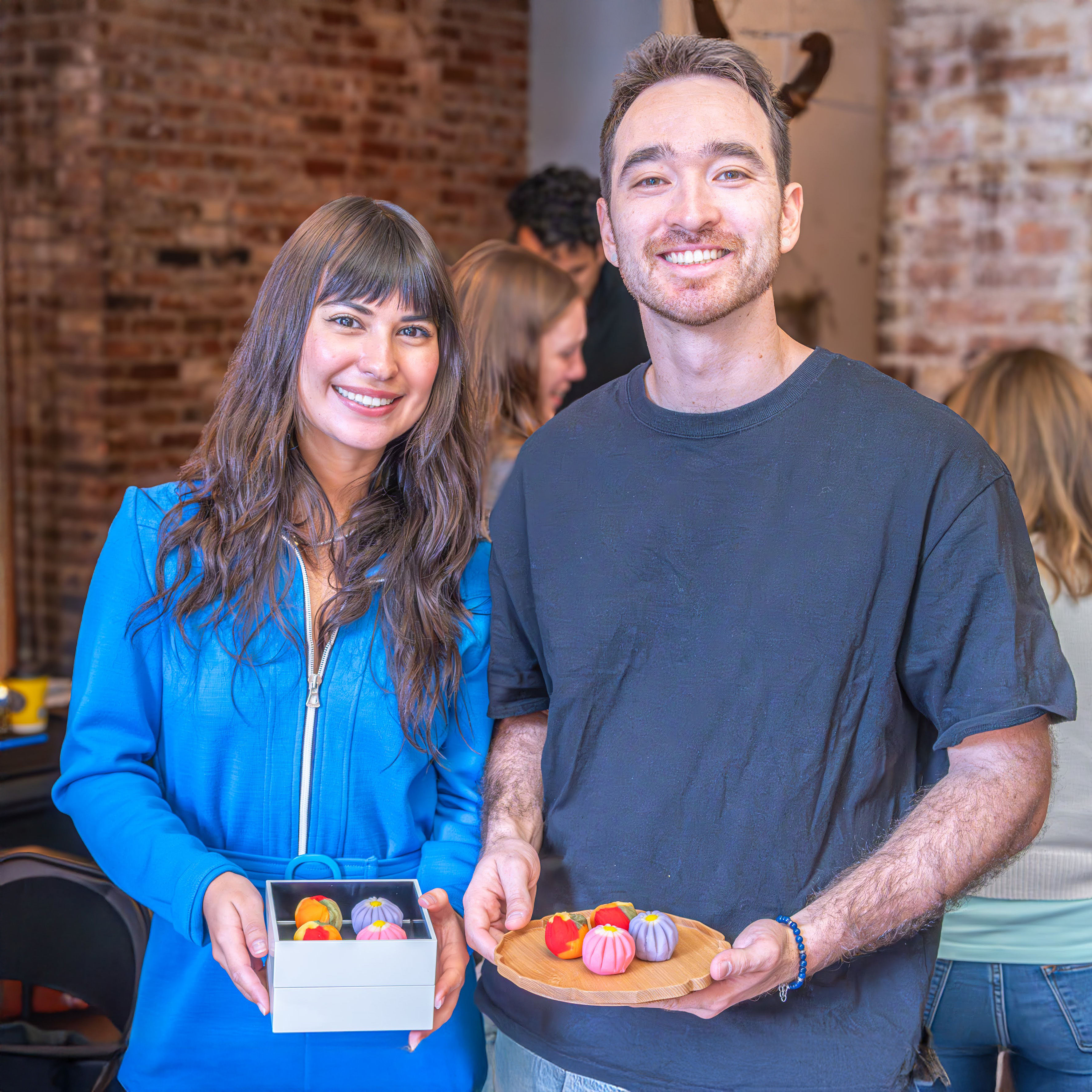 Smiling pair holding a box and wooden plate of colorful round sweets in a rustic brick-walled artisan pastry workshop.