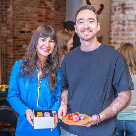 Smiling pair holding a box and wooden plate of colorful round sweets in a rustic brick-walled artisan pastry workshop.