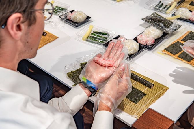 Participant wearing disposable gloves shaping pink sushi rice on a nori sheet over a bamboo rolling mat in an indoor sushi-making workshop with trays of fillings on a white table
