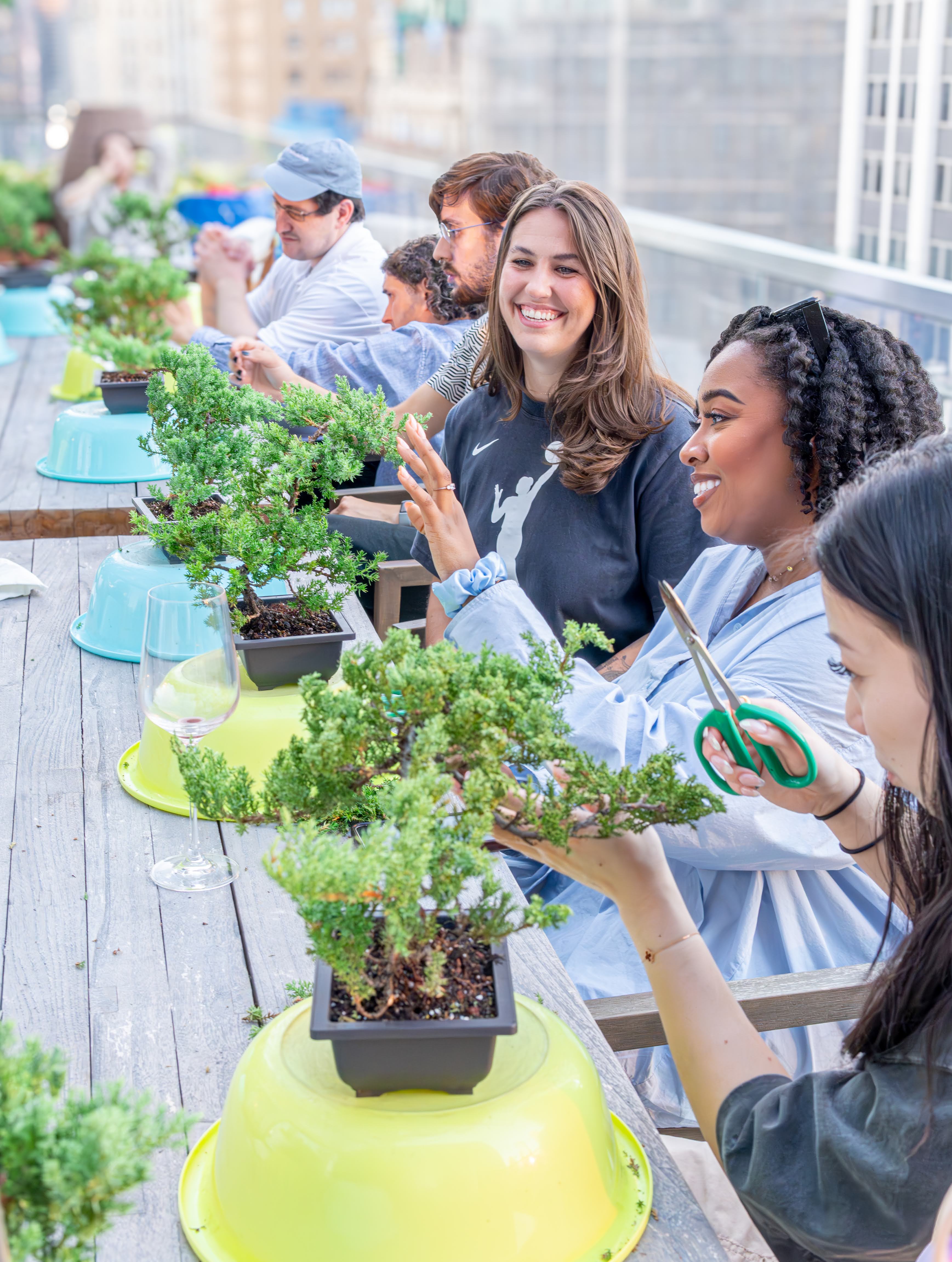 Smiling participants pruning small bonsai trees at a hands-on urban rooftop bonsai workshop with a city skyline in the background.