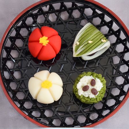 Four colorful wagashi (traditional Japanese confectionery) on a black lacquer woven tray with red rim: a red flower with yellow center, a white flower with yellow center, a green leaf-shaped sweet with gold flakes, and a green rice-style sweet topped with white paste and three azuki beans.