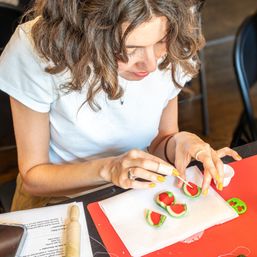 Person with curly hair decorating miniature green-and-red watermelon slices (clay or fondant) on a red craft mat