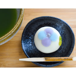 Top-down view of a delicate white Japanese wagashi decorated with pastel blue and purple flower, served on a black ceramic dish with a wooden pick and a bowl of green matcha tea on a bamboo tray.