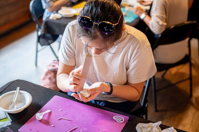 Focused crafter shaping a white clay ball with a sanding tool at an indoor craft workshop, sunglasses on head, purple work mat and bowl with utensils on the table