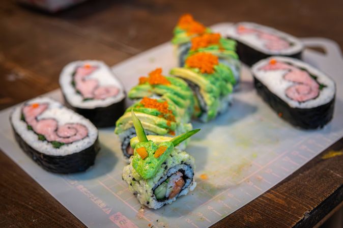 Close-up of colorful sushi: avocado-topped dragon roll with orange roe and green garnish, surrounded by sliced maki rolls on a silicone mat over a wooden table