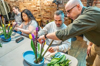 Hands-on flower-arranging workshop in a brick-walled urban studio — instructor guiding a participant arranging tulips and purple spike flowers in shallow blue vases.