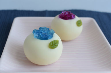 Two round white chocolate bonbons topped with blue and purple jelly crystals and small green leaf decorations on a pale pink ceramic plate