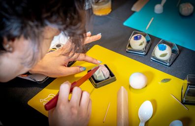 Hands sculpting a pastel pink wagashi (Japanese confection) with a red knife on a yellow cutting mat, surrounded by shaping tools, a wooden roller and flower-decorated sweets.