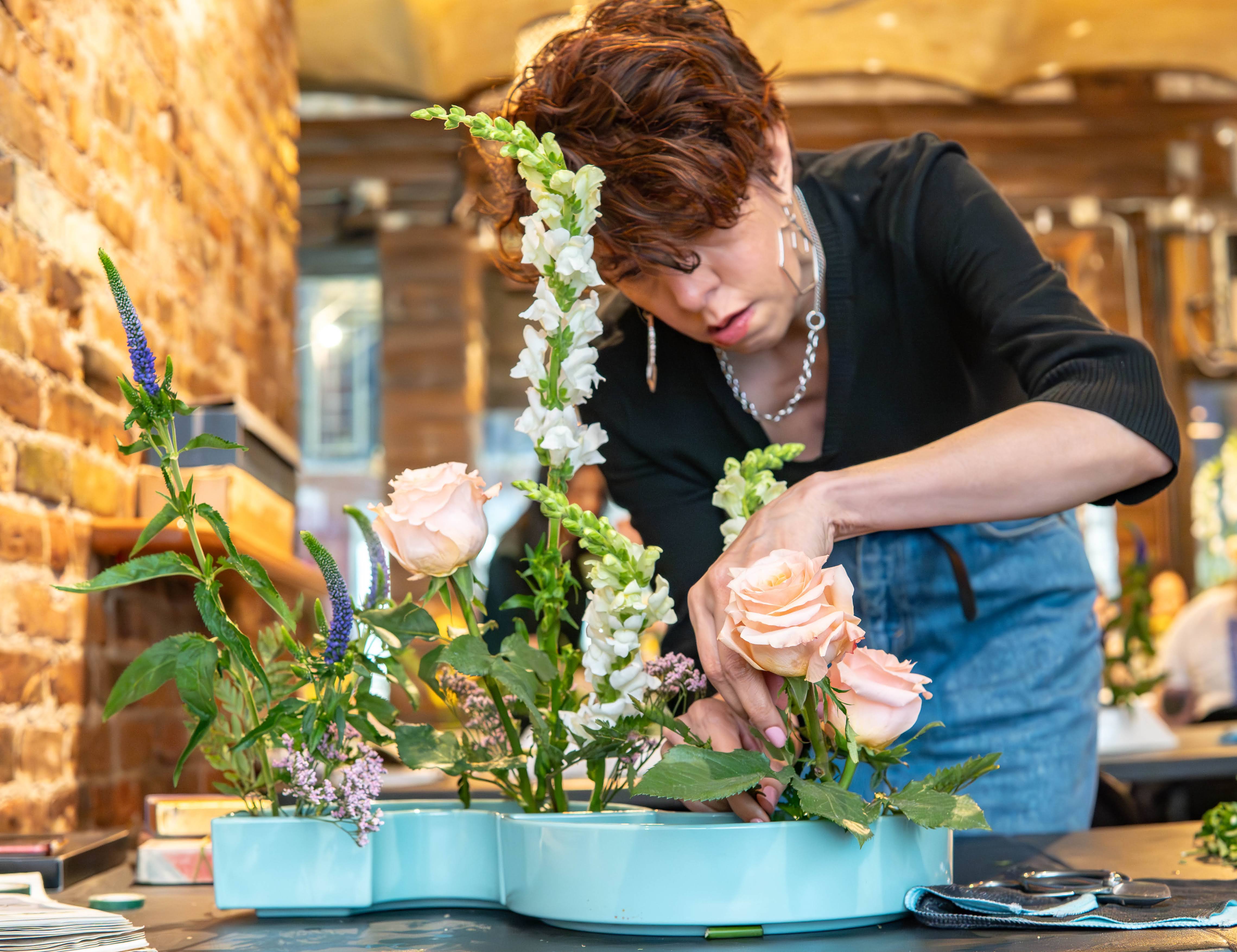 Close-up of a florist arranging peach roses, white snapdragons, and purple veronica in a turquoise ceramic planter on a table in a rustic brick-walled studio.