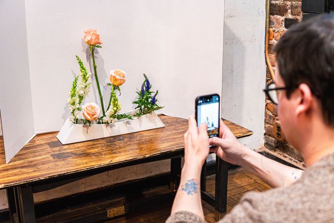 Person snapping a smartphone photo of a minimalist tabletop floral arrangement — peach roses, white snapdragons and purple veronica in a white geometric vase against a white backdrop on a wooden table with exposed brick wall.