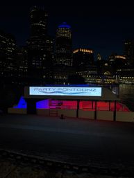Neon-lit party pontoon docked on a downtown harbor at night, blue and pink interior lights reflecting on the water with a dark city skyline and illuminated high-rises behind it.