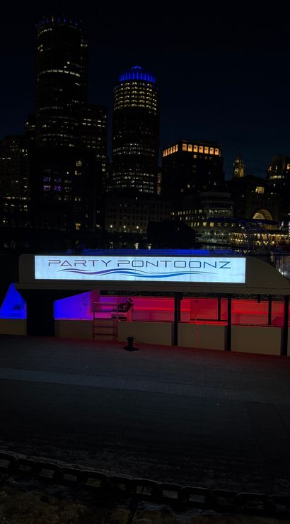 Neon-lit party pontoon docked on a downtown harbor at night, blue and pink interior lights reflecting on the water with a dark city skyline and illuminated high-rises behind it.