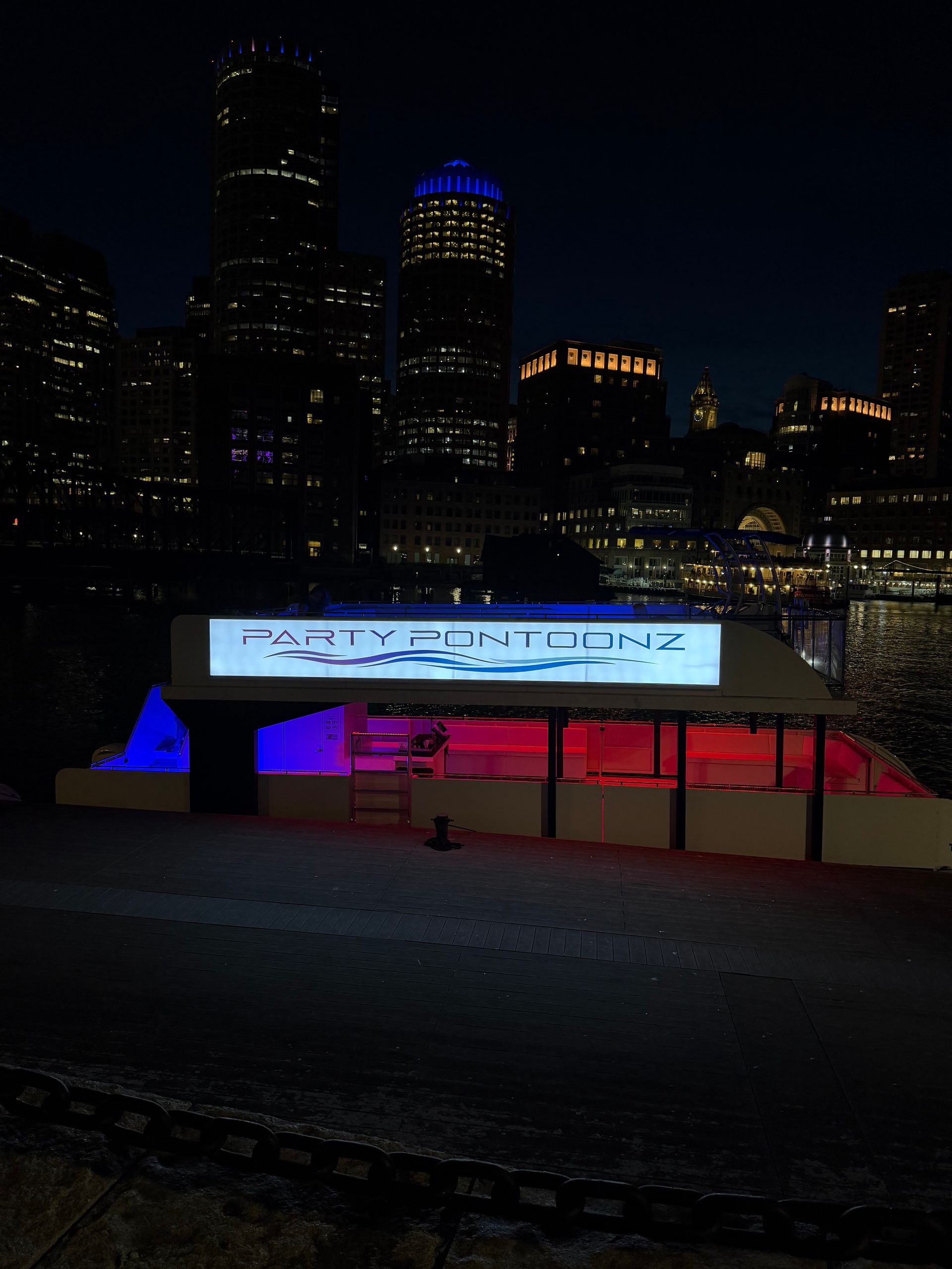 Neon-lit party pontoon docked on a downtown harbor at night, blue and pink interior lights reflecting on the water with a dark city skyline and illuminated high-rises behind it.