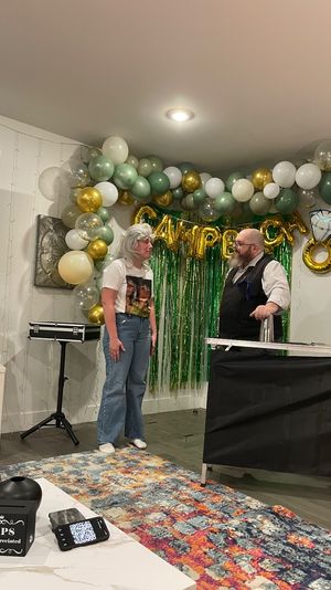 Living-room birthday party scene with green-and-gold balloon arch and foil fringe backdrop, gold HAPPY letter balloons, an entertainer speaking with a guest, and a TIP jar on the coffee table