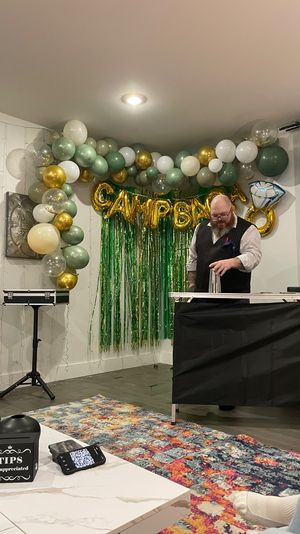 Party performer doing a cup-and-ball magic trick in a living room decorated with green, white and gold balloon garland and metallic green fringe backdrop; coffee table in foreground with a tips box, phone, and colorful rug.