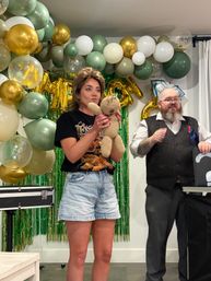 Indoor birthday party scene with a woman holding a knitted teddy bear while a bearded performer in a vest gestures beside her, gold, green and white balloon arch and green foil fringe backdrop with metallic letter balloons.