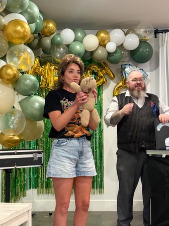 Indoor birthday party scene with a woman holding a knitted teddy bear while a bearded performer in a vest gestures beside her, gold, green and white balloon arch and green foil fringe backdrop with metallic letter balloons.