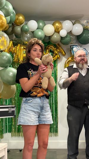 Indoor birthday party scene with a woman holding a knitted teddy bear while a bearded performer in a vest gestures beside her, gold, green and white balloon arch and green foil fringe backdrop with metallic letter balloons.