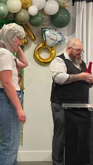 Smiling person wearing a white wig and veil watches a bearded magician in a vest perform a red sponge trick beneath green and gold balloon garland and an oversized ring balloon at a festive bachelorette-style party.