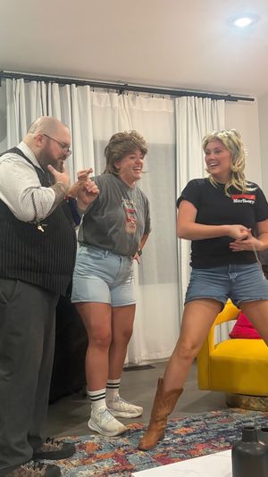 Three adults at a lively indoor house party — a man in glasses and vest and two women wearing wigs and denim shorts laugh and dance near a yellow armchair and balloon decorations.