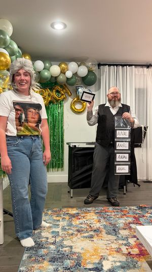 Two people at a home party in a living room with green, white and gold balloon arch and metallic fringe backdrop; one wears a gray wig and vintage-photo tee, the other holds a small “HUG” card and a vertical strip of labeled envelopes reading “ANAL” as part of a party game.