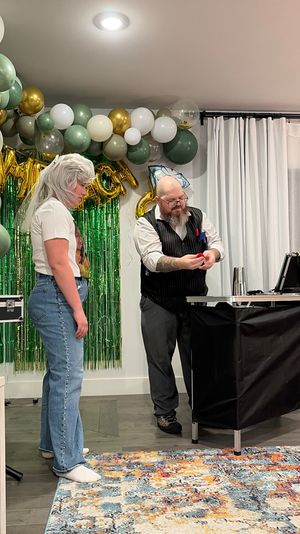 Indoor living-room party with a bearded magician in a vest performing a close-up trick at a table for a guest in a white wig and jeans, with green, white and gold balloon garland, green foil fringe backdrop and colorful area rug.
