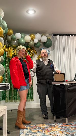 Festive indoor party scene with green, gold and white balloon garland and green fringe backdrop; a woman in a red jacket, denim shorts and brown cowboy boots stands beside a bearded man in a pinstripe vest and glasses by a table with party props.