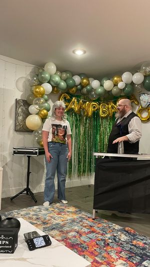 Home birthday party scene with green-and-gold balloon arch and tinsel backdrop, an adult in a graphic tee and wig standing beside a bearded performer in a vest doing a cup-and-ball magic trick, colorful area rug and a tip box on the coffee table.
