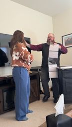 Bearded performer in a vest and glasses doing a card/magic trick with a vertical strip of pockets for a casually dressed woman in a cozy living room with a flat-screen TV and framed mountain photo.