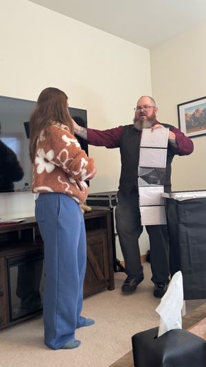 Bearded performer in a vest and glasses doing a card/magic trick with a vertical strip of pockets for a casually dressed woman in a cozy living room with a flat-screen TV and framed mountain photo.