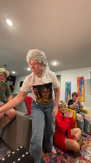 Group of friends in wigs at a cozy living-room costume party; one person reaches forward while others clap and sip wine, colorful paintings on the wall.