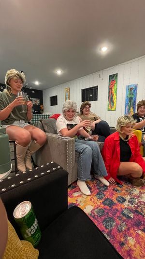 Group of five people in wigs laughing and holding wine glasses at a cozy indoor living-room gathering with white paneled walls, colorful abstract paintings, and a bright patterned rug.
