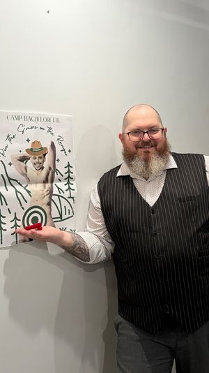 Smiling bearded man in glasses wearing a pinstriped vest indoors, holding a red dart in front of a 'Camp Bachelorette' party-game poster featuring a playful shirtless cutout with a target.