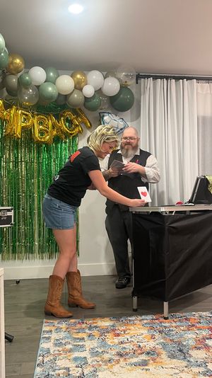 Magician performing a card trick at an indoor party: bearded man in a vest fans cards while a woman in denim shorts and cowboy boots reveals a card with a red heart under green fringe and gold and white balloons.