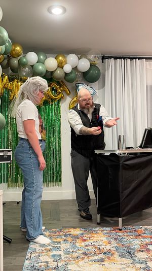 Bearded magician performing a close-up trick with a small red ball for a standing guest in front of a green-and-gold balloon garland and fringe backdrop at an indoor party.